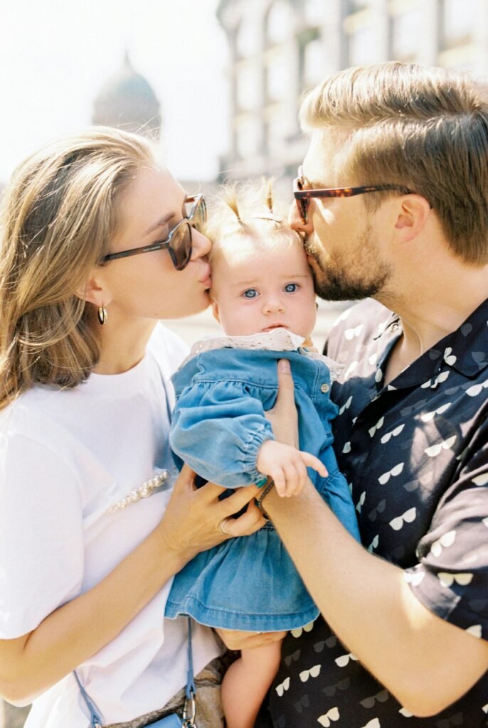 A joyful family portrait with parents kissing their baby outdoors on a sunny day.